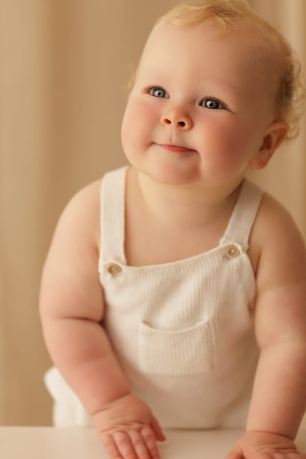 A baby in cream dungarees smiling on a warm backdrop