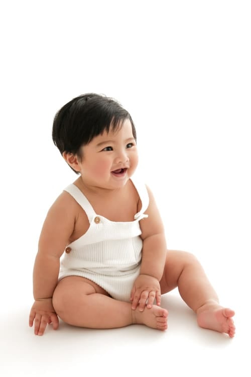 Baby sitting upright in a cream romper on a white backdrop