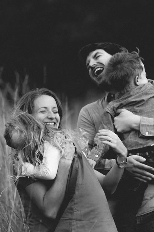 A family laughing together in tall grass at dusk, a child carried on his father's shoulder