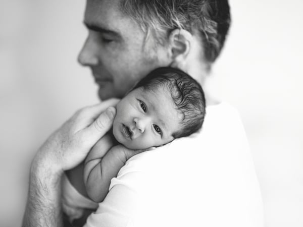 A father holding his newborn against his shoulder, the baby awake and looking towards the camera, in black and white