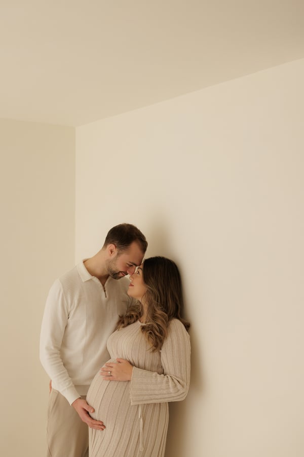 A couple standing together in a pale corner, both dressed in cream, hands on her bump