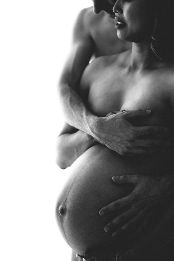 A partner standing behind with both arms wrapped around a bare pregnant belly, in black and white