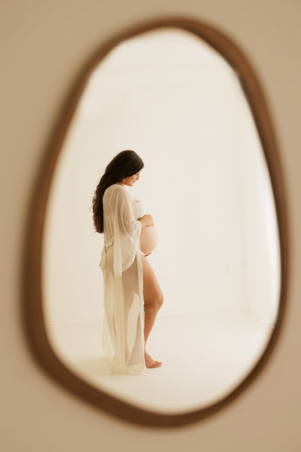 An expectant mother in a cream gown reflected in an oval mirror