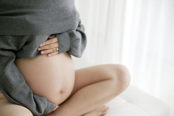 An expectant mother seated against a pale backdrop, a grey jumper lifted over her bump