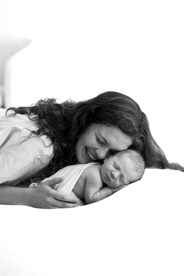 A mother lying beside her swaddled newborn on a white bed, in black and white