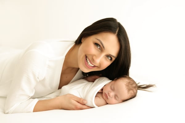 A mother lying beside her swaddled newborn on a white backdrop, smiling towards the camera