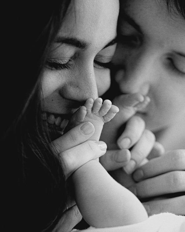 Close-up in black and white of a smiling mother with her newborn's foot resting against her face