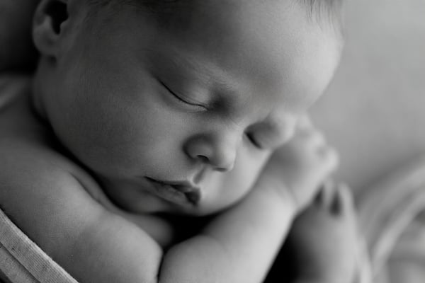 Close-up of a newborn asleep with both hands tucked near the face, in black and white
