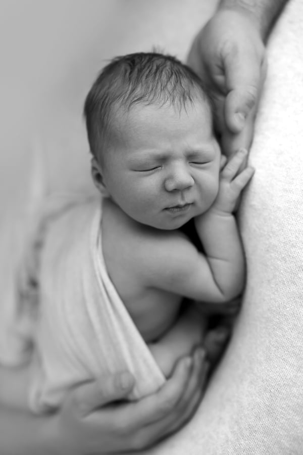 A newborn asleep in a wrap, a hand supporting their head on a textured blanket, in black and white
