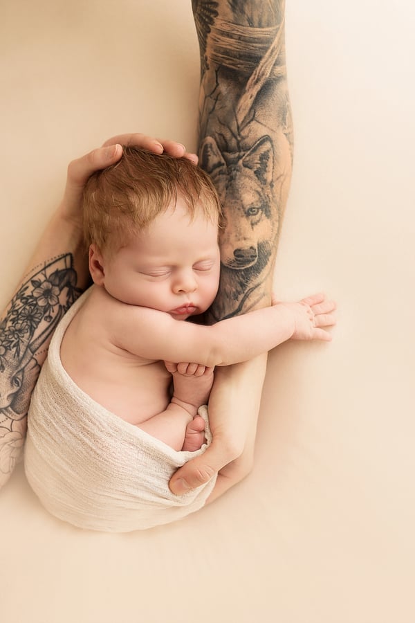 Newborn asleep in a cream wrap on a cream backdrop, cradled by a heavily tattooed arm