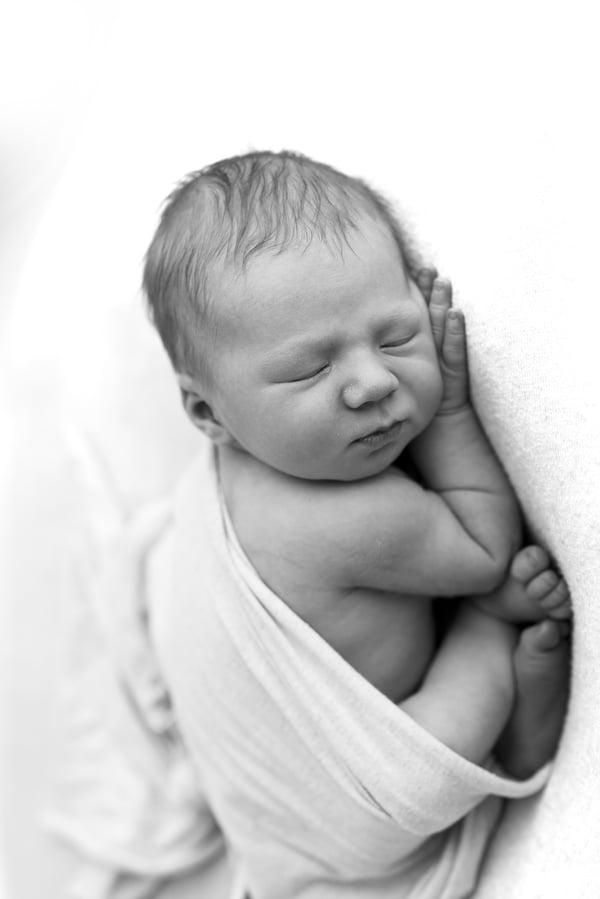 A newborn curled asleep in a white wrap on a white backdrop, in black and white