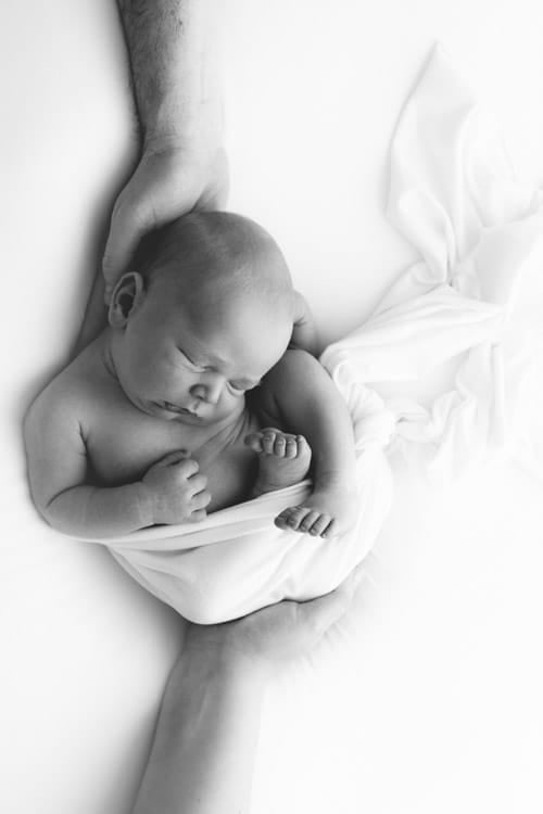 Newborn lying on its back on a white wrap, head resting in a parent's hand