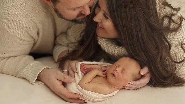 Parents lying either side of their swaddled newborn, foreheads close together, smiling