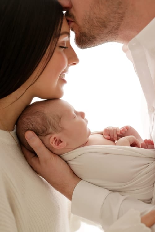 Two parents leaning their foreheads together over their swaddled newborn
