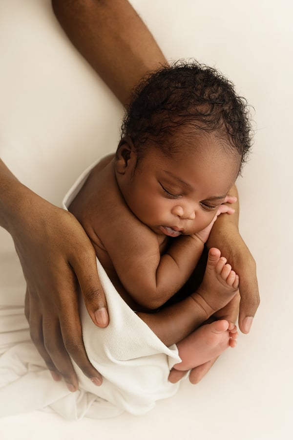 Newborn with dark curly hair asleep, cradled in a parent's hands on a white backdrop