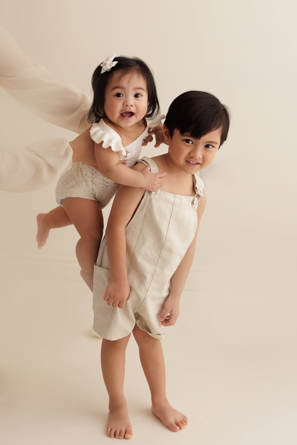 An older child carrying their younger sibling on one hip in the studio, both in cream