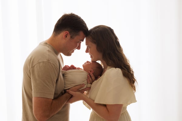 Both parents standing close together holding their newborn, backlit by a bright window