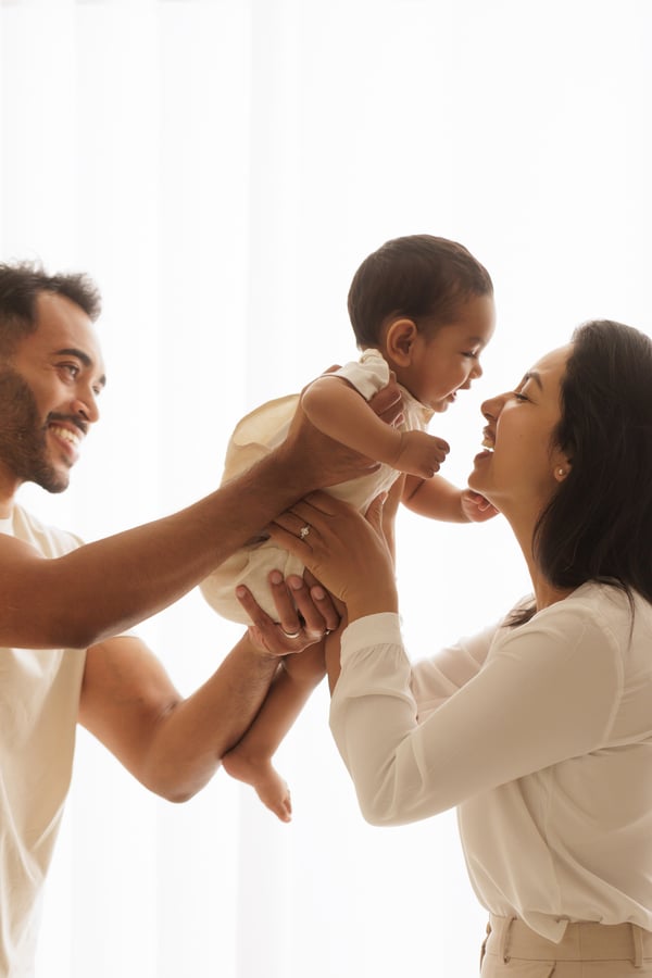 Both parents lifting their baby up between them against a bright window, all three laughing