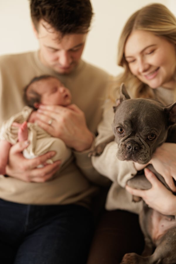 Parents sitting together, one holding their swaddled newborn and the other holding the family's French bulldog