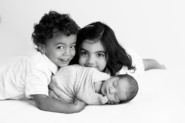 An older brother and sister lying down together with their swaddled newborn sibling, in black and white